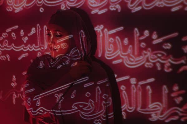 A woman standing against a wall with Arabic text projected over her.